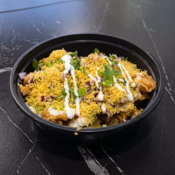 A black bowl of Indian chaat, topped with sev, chopped red onions, cilantro, and white sauce.