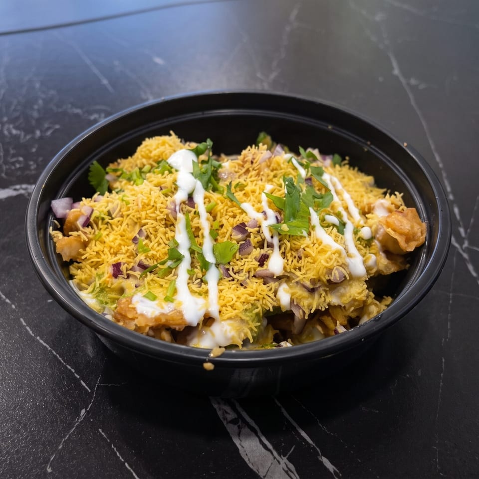 A black bowl of Indian chaat, topped with sev, chopped red onions, cilantro, and white sauce.