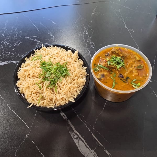 A black bowl of rice with herbs and a plastic container of curry on a dark marbled table.